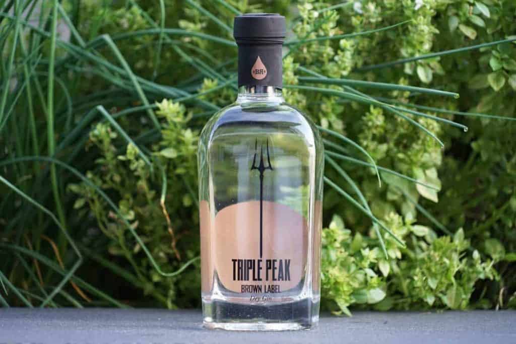Im Freien steht eine durchsichtige Flasche mit der Aufschrift „Triple Peak Brown Label“, umgeben von grünen Pflanzen. Auf der Flasche ist ein schwarzes Dreizack-Logo zu sehen, und am unteren Rand befindet sich ein braunes Etikett.