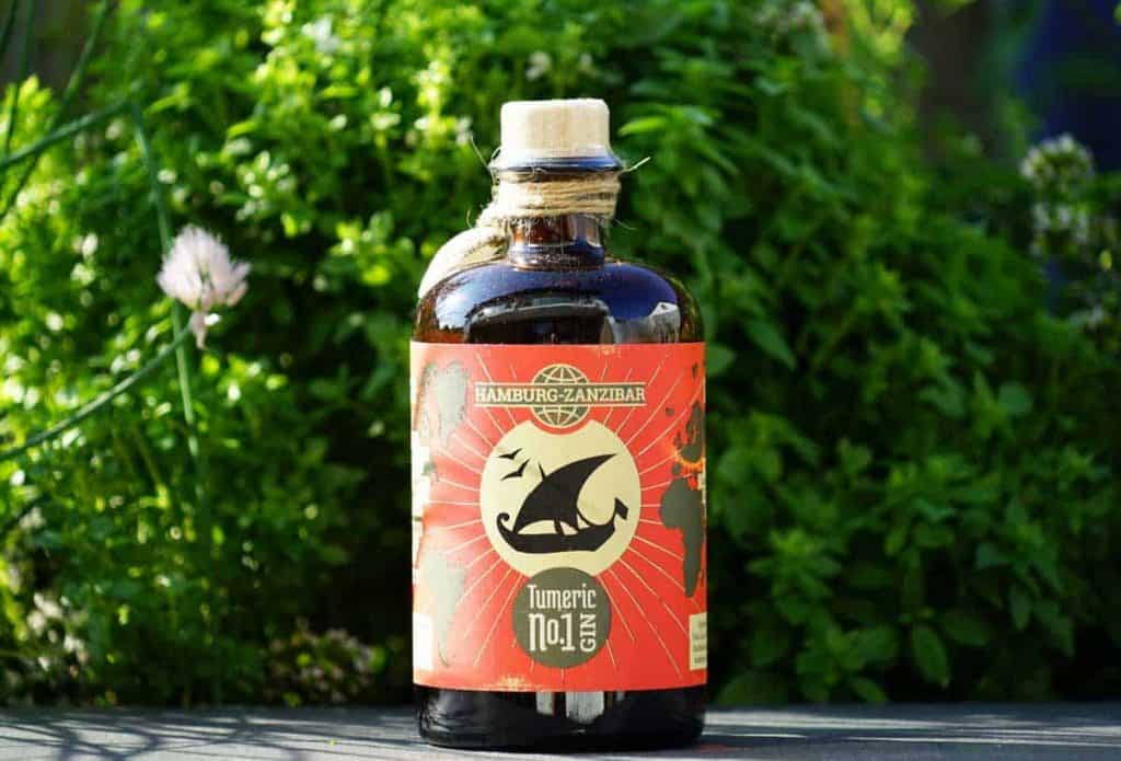 Auf einer Fläche steht eine braune Glasflasche mit der Aufschrift „Hamburg-Zanzibar Turmeric No. 1 Gin“ und einer schwarzen Tiersilhouette, umgeben von grünem Laub und Pflanzen im Hintergrund.