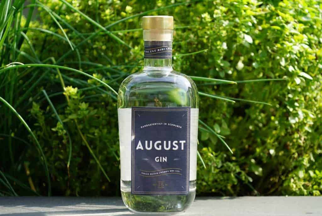 Eine Flasche „August Gin“ mit dunklem Etikett steht im Freien auf einer grauen Fläche, umgeben von grünen, üppig blättrigen Pflanzen in hellem Tageslicht.