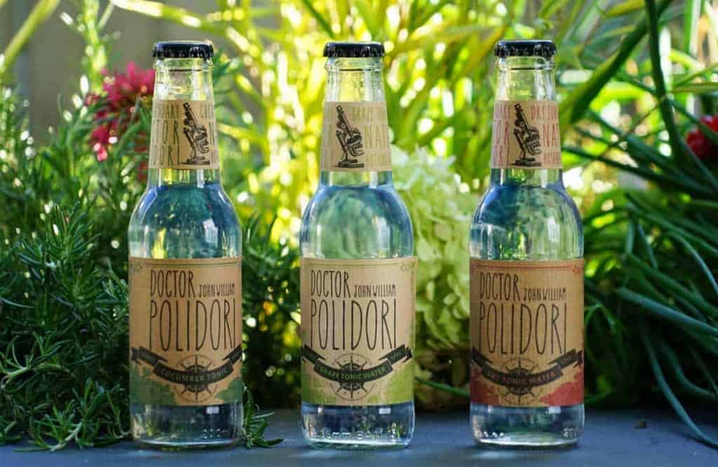 Drei Glasflaschen mit „Doctor Polidori“-Tonic Water stehen auf einem Tisch im Freien, umgeben von grünen Pflanzen und bunten Blumen im Hintergrund.