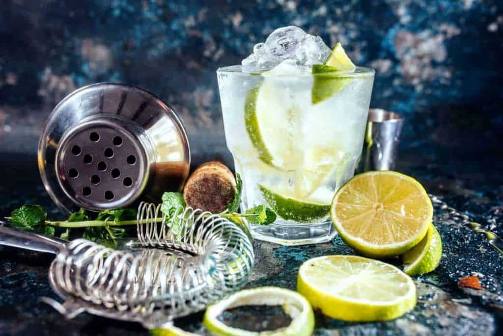 Auf einer dunklen, strukturierten Oberfläche stehen ein Cocktailglas mit Eis und Limettenschnitzen neben einem Cocktailshaker, einem Sieb, einem Muddler, Minzblättern und Limettenscheiben.