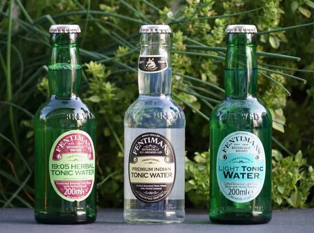 Drei Flaschen Fentimans Tonic Water – „Herbal“, „Premium Indian“ und „Light“ – stehen im Freien nebeneinander, im Hintergrund sind grüne Pflanzen verschwommen zu sehen.