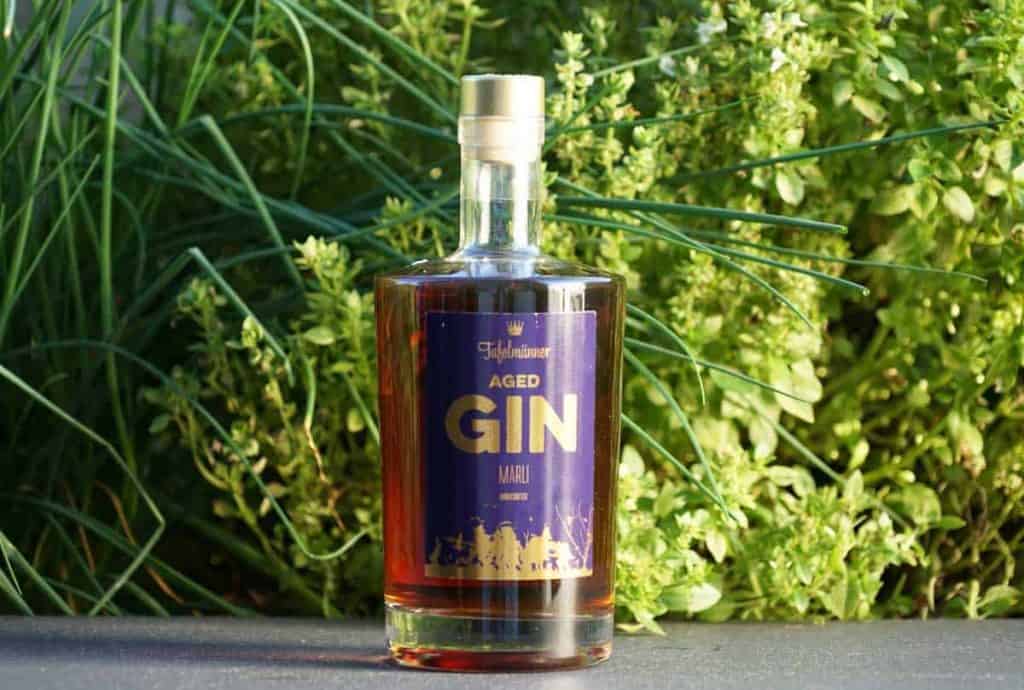 Eine Flasche „Highclere Aged Gin“ mit einem violett-goldenen Etikett steht auf einer ebenen Fläche, im Hintergrund sind grüne Pflanzen und Blätter zu sehen.