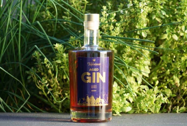Eine Flasche „Highclere Aged Gin“ mit einem violett-goldenen Etikett steht auf einer ebenen Fläche, im Hintergrund sind grüne Pflanzen und Blätter zu sehen.