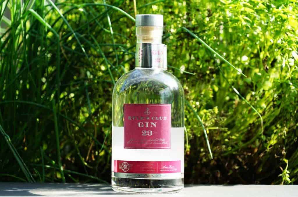Eine Flasche „Rivet Club Gin 23“ mit rotem Etikett steht im Freien auf einer Fläche, im Hintergrund ist sie von grünem Laub umgeben.