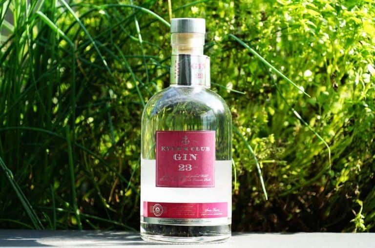 Eine Flasche „Rivet Club Gin 23“ mit rotem Etikett steht im Freien auf einer Fläche, im Hintergrund ist sie von grünem Laub umgeben.