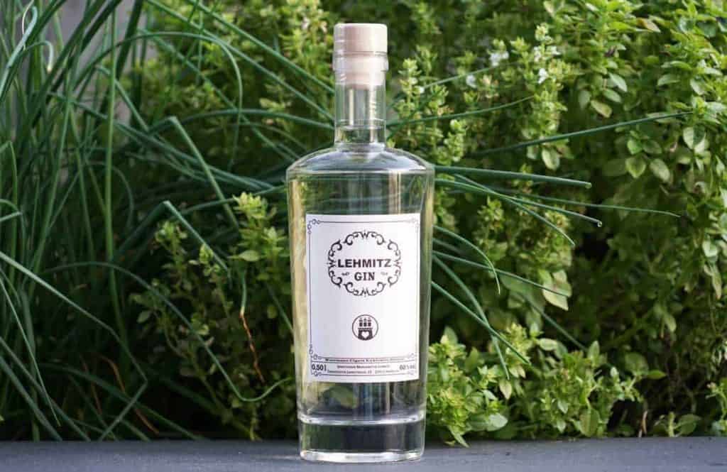 Vor üppig grünen Kräutern und Pflanzen steht eine klare Glasflasche mit Elemitz Gin und einem weißen Etikett.