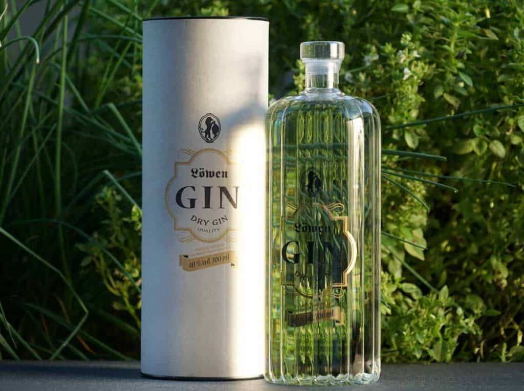 Eine durchsichtige, gerippte Flasche „Löwen Dry Gin“ steht neben ihrer zylindrischen weißen Verpackung; beide sind im Freien vor dem Hintergrund grüner Pflanzen zu sehen.