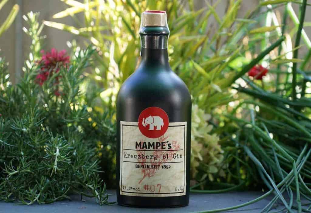 Eine dunkelgrüne Flasche „Mampes Kreuzberg 61 Gin“ mit einem Etikett, auf dem ein rotes Elefantenlogo abgebildet ist, steht im Freien inmitten von grünen Pflanzen und Blumen.