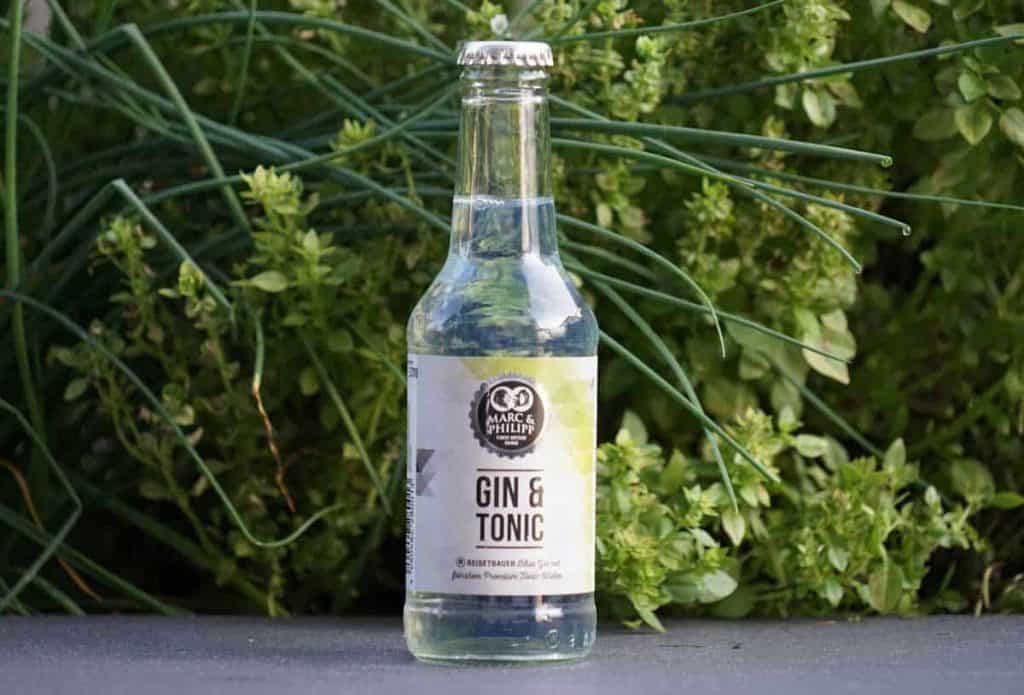 Auf einer dunklen Fläche steht eine durchsichtige Flasche mit der Aufschrift „Marc & Philipp Gin & Tonic“, im Hintergrund sind grüne Blattpflanzen zu sehen.