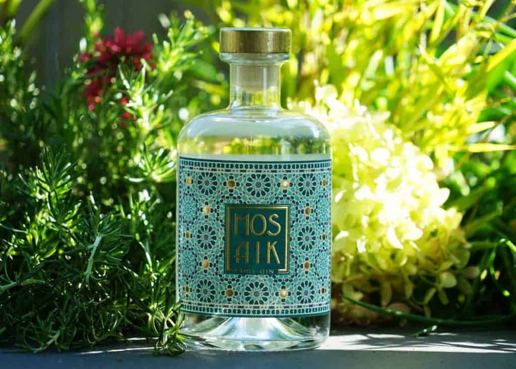 Eine durchsichtige Flasche mit der Aufschrift „Mosaik Dry Gin“ und einem blau-goldenen Mosaikmuster steht im Freien inmitten von grünen Kräutern und gelben Blumen im strahlenden Sonnenlicht.