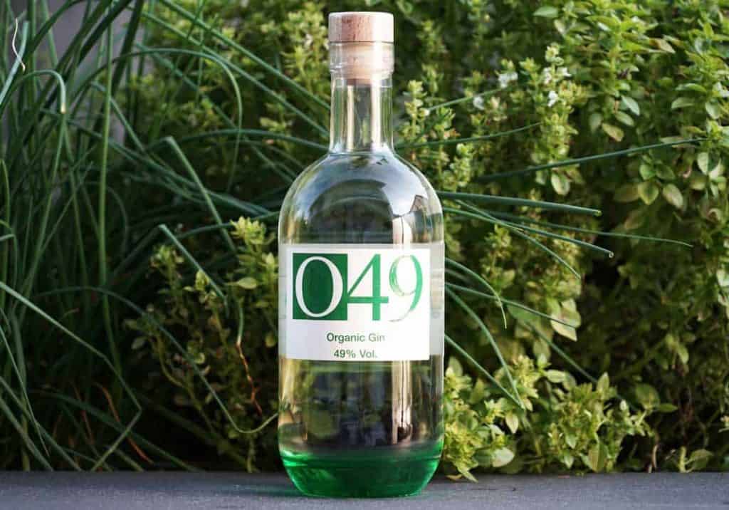 Eine klare Glasflasche mit der Aufschrift „049 Organic Gin 49 % Vol.“ und einem Korkverschluss, die vor üppig grünen Kräutern und Pflanzen steht.