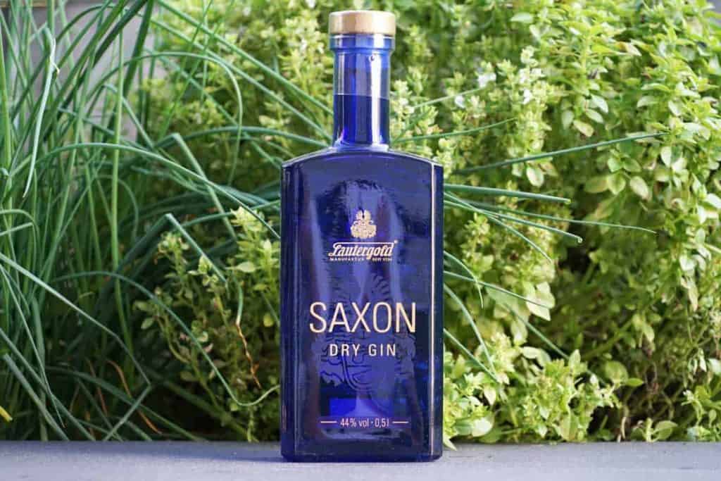 Eine blaue Glasflasche „Saxon Dry Gin“ steht im Freien auf einer Fläche, im Hintergrund sind grüne Pflanzen und Blätter zu sehen.