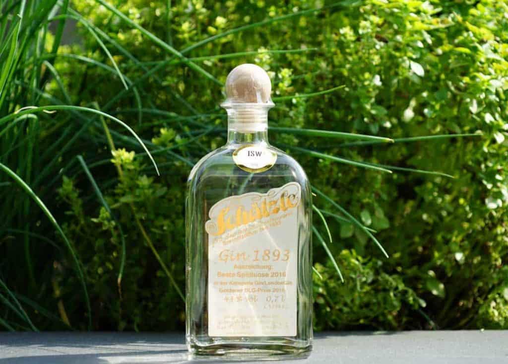 Eine Flasche „Schlichte Gin 1893“ mit einem Holzkorken steht im Freien auf einer dunklen Oberfläche, im Hintergrund sind grüne, blättrige Pflanzen zu sehen. Das Etikett ist gold-weiß und enthält Angaben zu Auszeichnungen.
