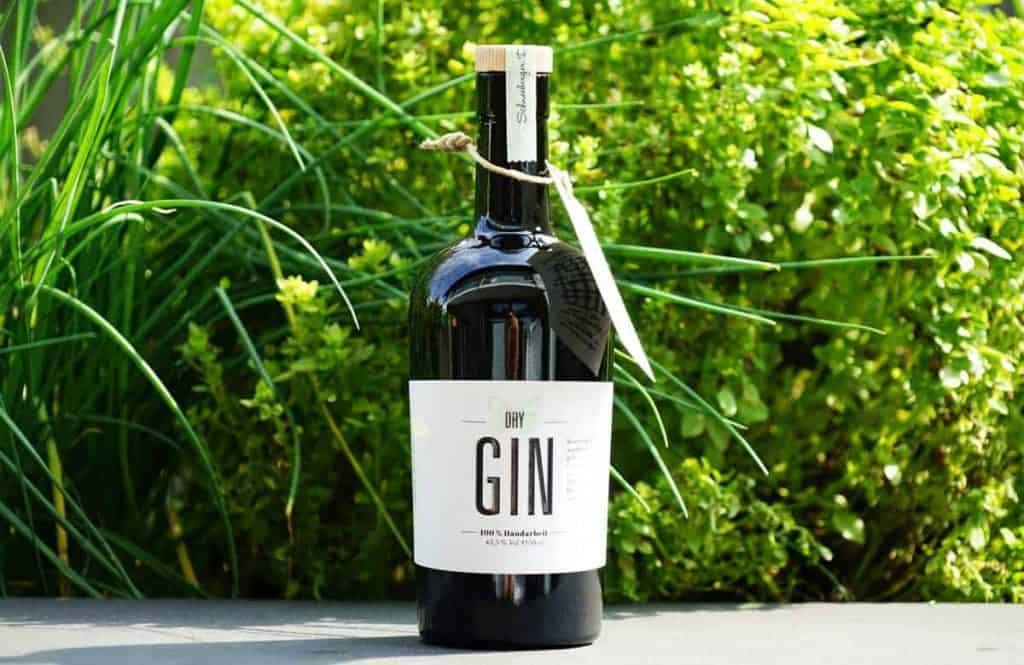 Auf einer grauen Fläche steht eine dunkle Glasflasche mit trockenem Gin, die mit einem weißen Etikett und einem Anhänger versehen ist; im Hintergrund sind üppig grüne Pflanzen zu sehen.