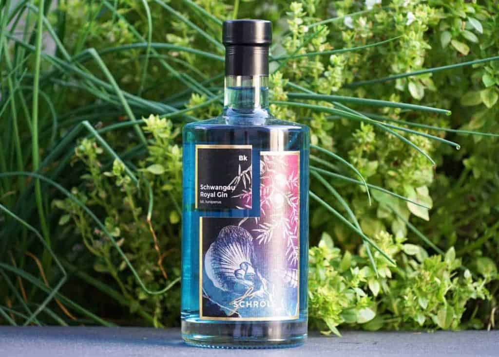 Eine durchsichtige Flasche „Schwangau Royal Gin“ mit einem schwarzen, blumigen Etikett steht im Freien auf einer Fläche, im Hintergrund umgeben von üppig grünen Pflanzen.