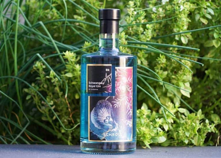 Eine durchsichtige Flasche „Schwangau Royal Gin“ mit einem schwarzen, blumigen Etikett steht im Freien auf einer Fläche, im Hintergrund umgeben von üppig grünen Pflanzen.