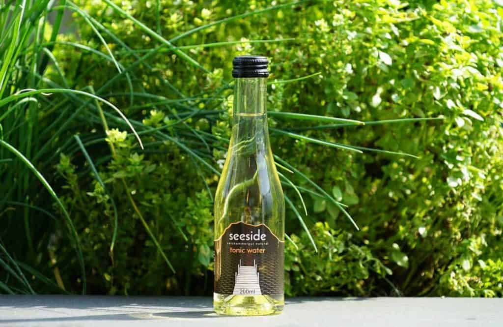 Eine durchsichtige Flasche Seeside Tonic Water mit schwarzem Verschluss steht auf einer grauen Fläche, im Hintergrund sind grüne Blattpflanzen zu sehen.