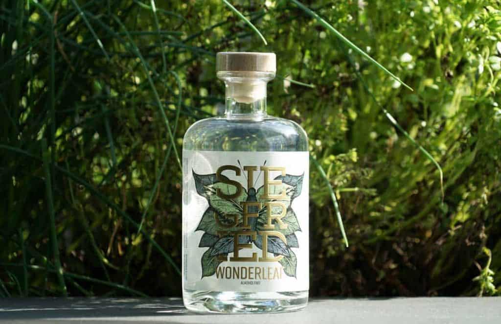 Auf einer dunklen Fläche steht eine durchsichtige Glasflasche mit der Aufschrift „Siegered Wonderleaf“, im Hintergrund sind üppig grüne Pflanzen zu sehen. Die Flasche hat einen Holzverschluss und ein Etikett mit botanischem Motiv.
