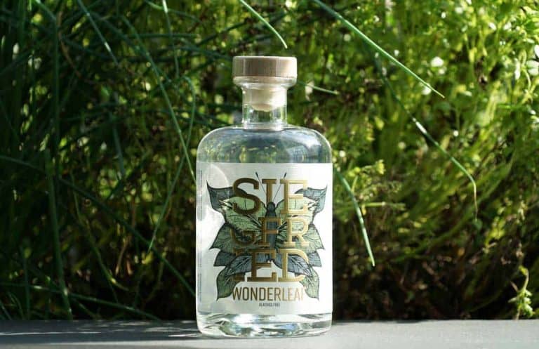 Auf einer dunklen Fläche steht eine durchsichtige Glasflasche mit der Aufschrift „Siegered Wonderleaf“, im Hintergrund sind üppig grüne Pflanzen zu sehen. Die Flasche hat einen Holzverschluss und ein Etikett mit botanischem Motiv.