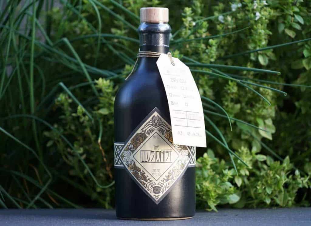 Eine schwarze Flasche mit der Aufschrift „The Illusionist“, einem Holzkorken und einem Papieranhänger, die im Freien vor grünen Pflanzen steht.
