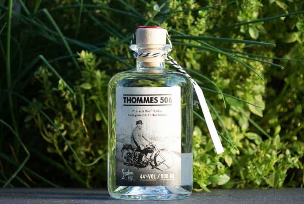 Eine durchsichtige Flasche mit der Aufschrift „THOMMES 506“ und der Abbildung einer Person auf einem Motorrad, die im Freien vor grünem Laub sitzt. Auf dem Etikett sind 44 % Vol. und ein Fassungsvermögen von 500 ml angegeben.