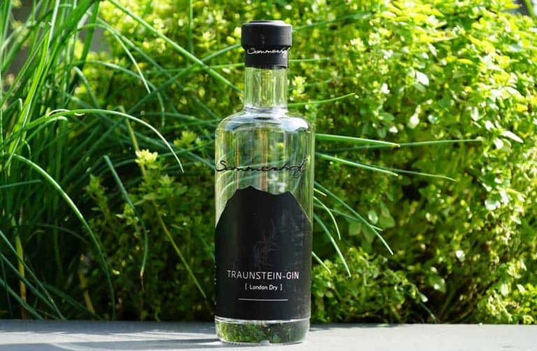 Eine durchsichtige Flasche „Traunstein-Gin London Dry“ steht im Freien auf einer Fläche, umgeben von üppig grünen Pflanzen und Gras im Hintergrund.