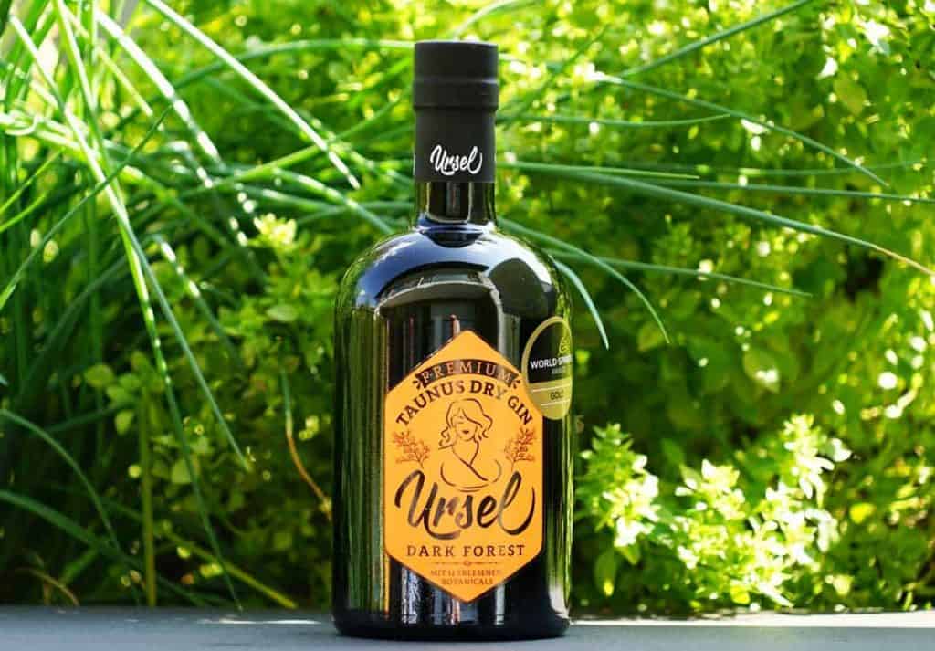 Eine schwarze Flasche „Ursel Taunus Dry Gin“ mit gelbem Etikett steht draußen auf einer Fläche, im Hintergrund umgeben von leuchtend grünen Pflanzen.