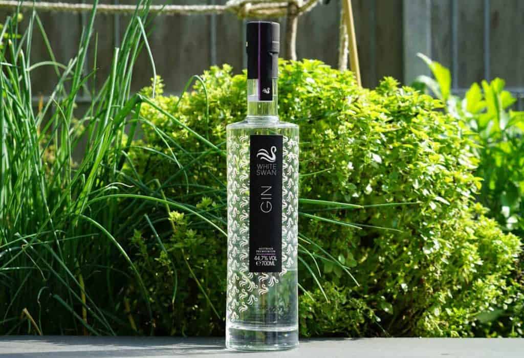 Eine Flasche „White Swan Gin“ steht im Freien auf einer dunklen Fläche, umgeben von grünen Blattpflanzen im strahlenden Sonnenlicht.