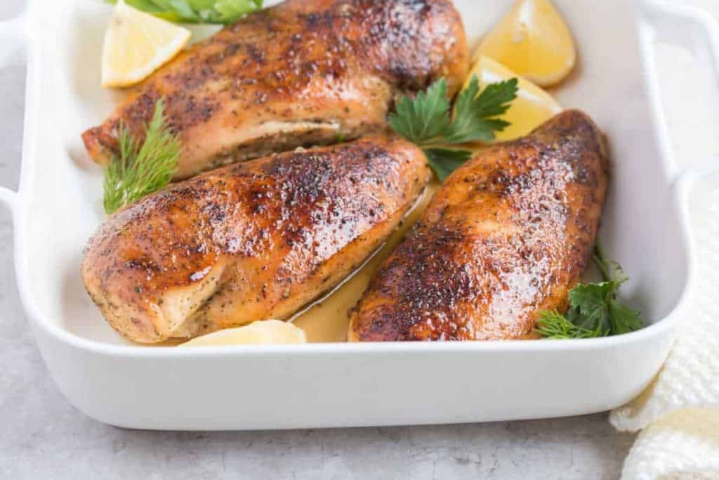 Drei gebratene Hähnchenbrustfilets mit goldbrauner Kruste werden in einer weißen Auflaufform serviert, garniert mit Zitronenschnitzen und frischen Kräutern.