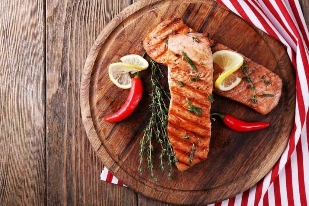 Gegrillte Lachsfilets auf einer runden Holzplatte, garniert mit Zitronenscheiben, frischem Thymian und roten Chilischoten, serviert auf einem rustikalen Holztisch mit einer rot-weiß gestreiften Tischdecke.