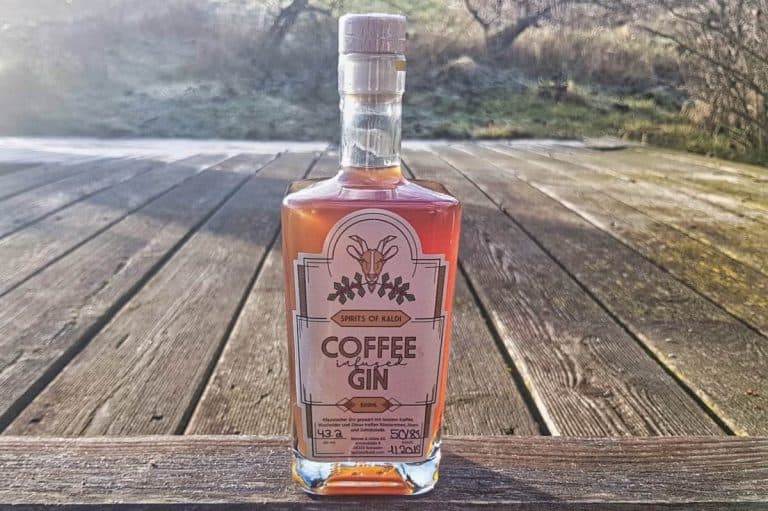 Auf einer Holzterrasse im Freien steht eine Flasche mit Kaffee aromatisiertem Gin; im unscharfen Hintergrund sind Bäume und Gras zu sehen.