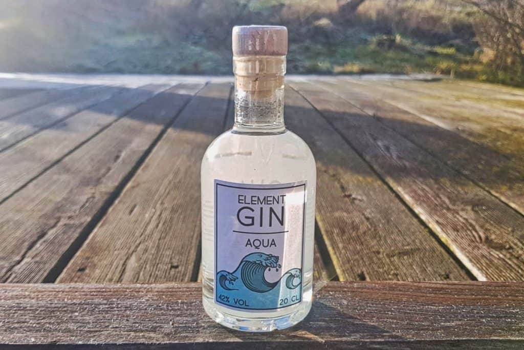 Eine klare Glasflasche „Element Gin Aqua“ steht auf einer Holzterrasse im Freien, im Hintergrund sind Sonnenlicht und Natur zu sehen.