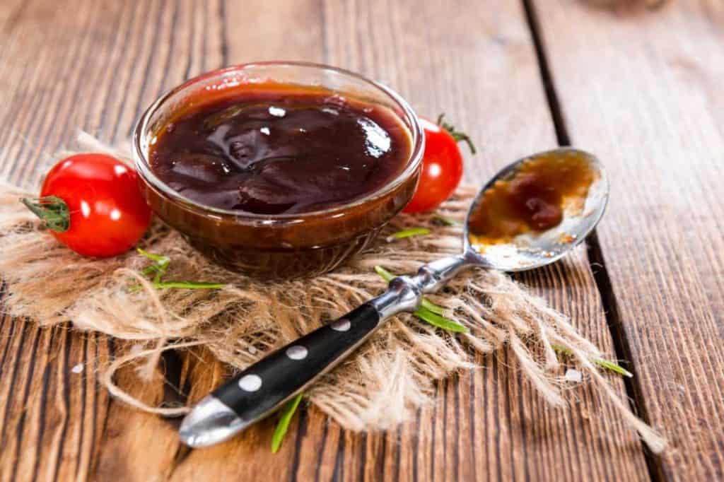 Auf einem rustikalen Holztisch steht eine Glasschüssel mit Barbecue-Sauce auf einem Stück Sackleinen, daneben liegen ein Löffel mit Sauce und zwei kleine Tomaten.