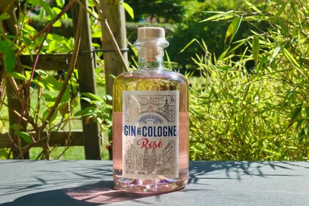 Auf einem schwarzen Tisch im Freien steht eine Flasche „Gin de Cologne Rosé“, umgeben von grünen Pflanzen und im Sonnenlicht.