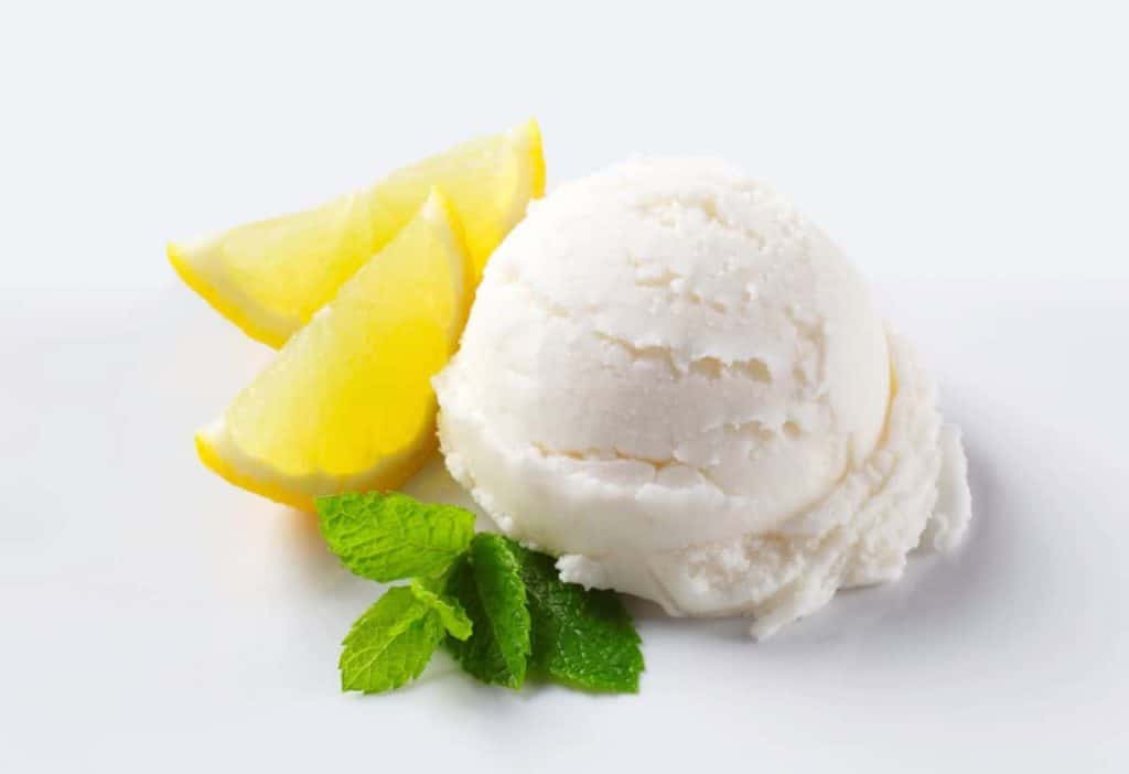 Eine Kugel Zitronensorbet wird auf einem weißen Teller mit zwei Zitronenschnitzen und einem Zweig frischer Minze serviert.