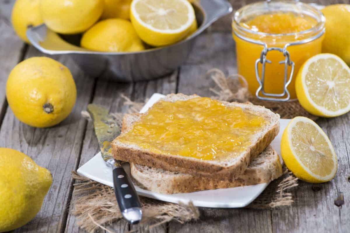 Zwei Scheiben Brot mit Zitronenmarmelade auf einem weißen Teller, umgeben von frischen Zitronen, einem Glas Zitronenmarmelade, einem Buttermesser und einer Metallschüssel mit Zitronen auf einem rustikalen Holztisch.