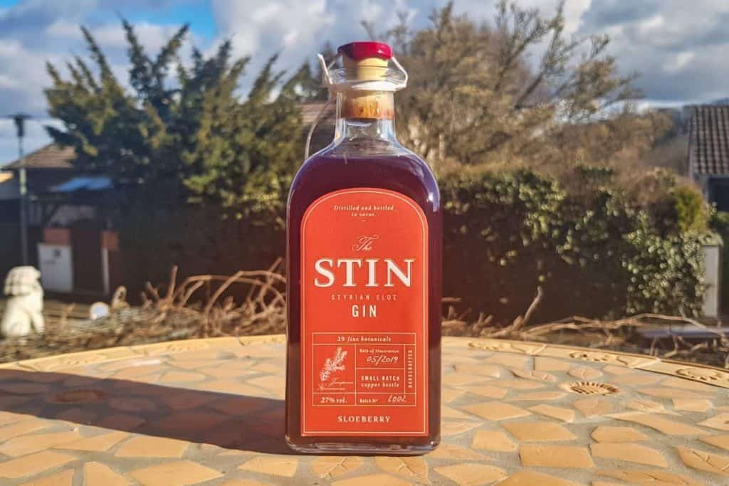 Auf einem Mosaiktisch im Freien steht eine Flasche STIN Steirischer Schlehen-Gin, im Hintergrund sind Bäume, Häuser und ein teilweise bewölkter Himmel zu sehen.