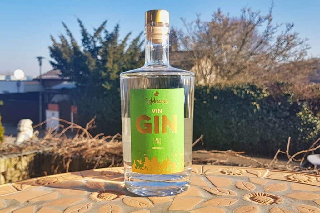 Auf einem Mosaiktisch im Freien steht eine Gin-Flasche aus klarem Glas mit einem grün-gelben Etikett; im Hintergrund sind unter blauem Himmel Bäume und ein Haus zu sehen.