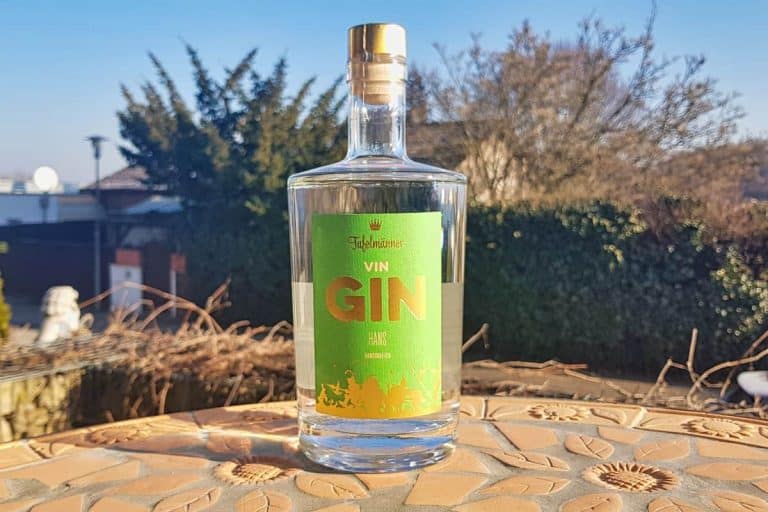Auf einem Mosaiktisch im Freien steht eine Gin-Flasche aus klarem Glas mit einem grün-gelben Etikett; im Hintergrund sind unter blauem Himmel Bäume und ein Haus zu sehen.