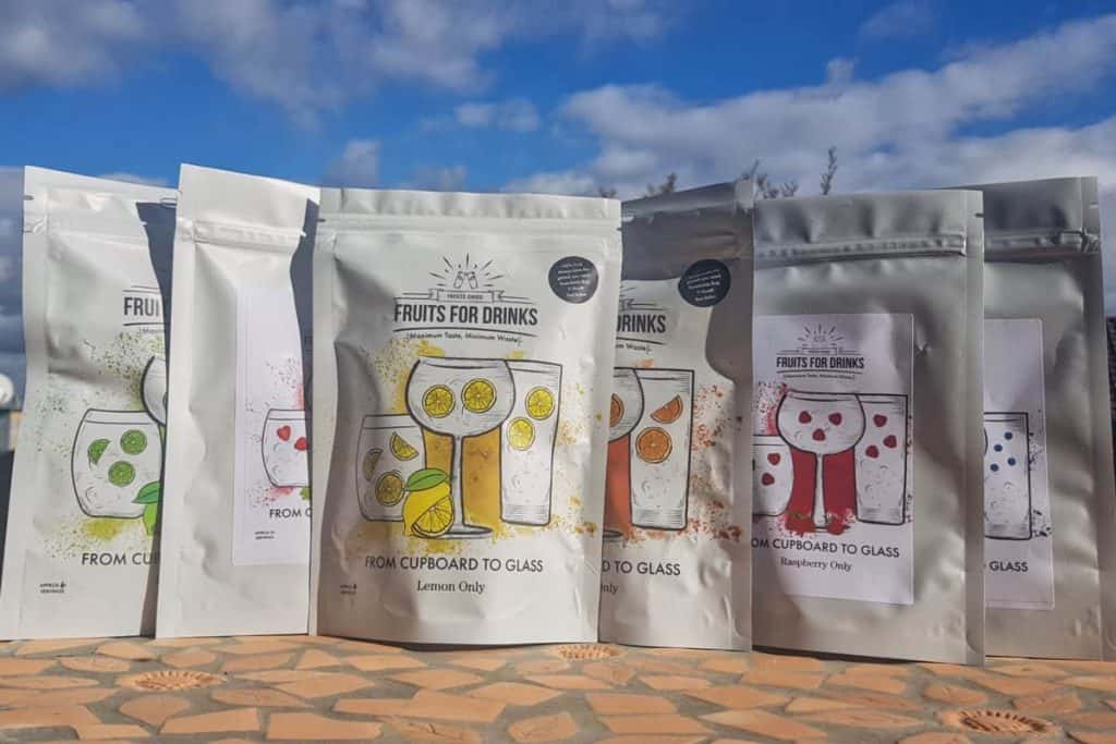 Mehrere bunte Packungen „Fruits for Drinks“ sind im Freien auf einer gemusterten Fläche unter blauem Himmel ausgestellt; jede davon ist mit Abbildungen von Früchten und Gläsern verziert, darunter die Geschmacksrichtungen Zitrone und Himbeere.