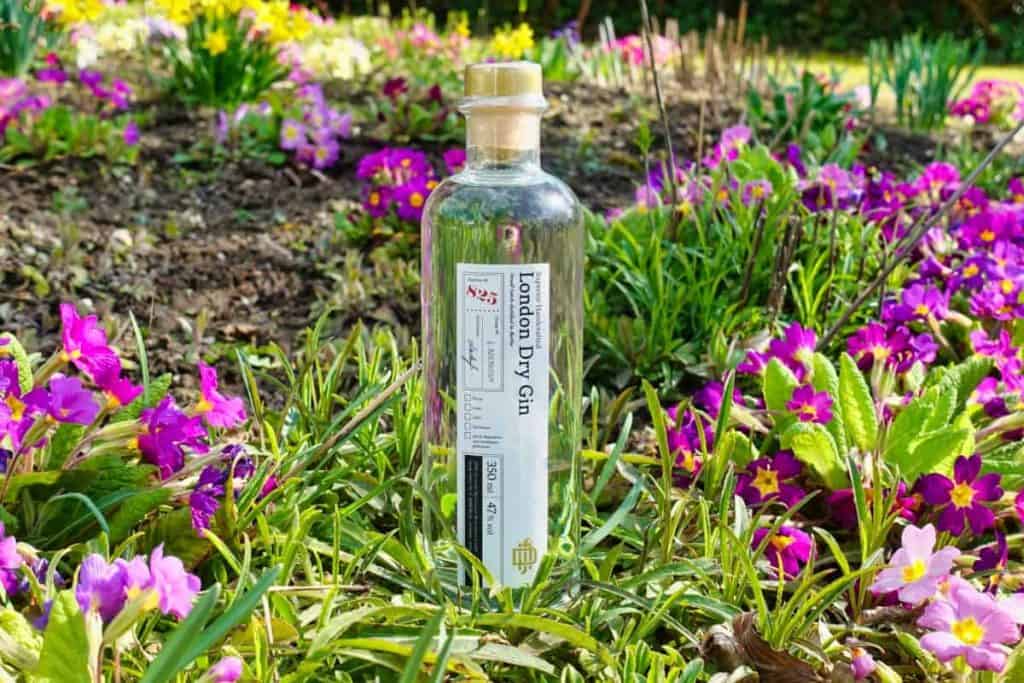 Eine durchsichtige Flasche London Dry Gin steht aufrecht inmitten von grünem Gras und leuchtend rosa und gelben Blumen in einer Gartenkulisse.
