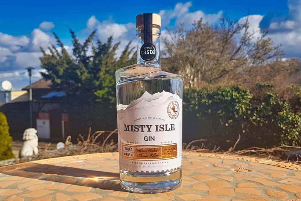 Eine Flasche Misty Isle Gin steht auf einem sonnenbeschienenen Tisch im Freien, im Hintergrund sind Bäume, Sträucher und ein blauer Himmel mit Wolken zu sehen.