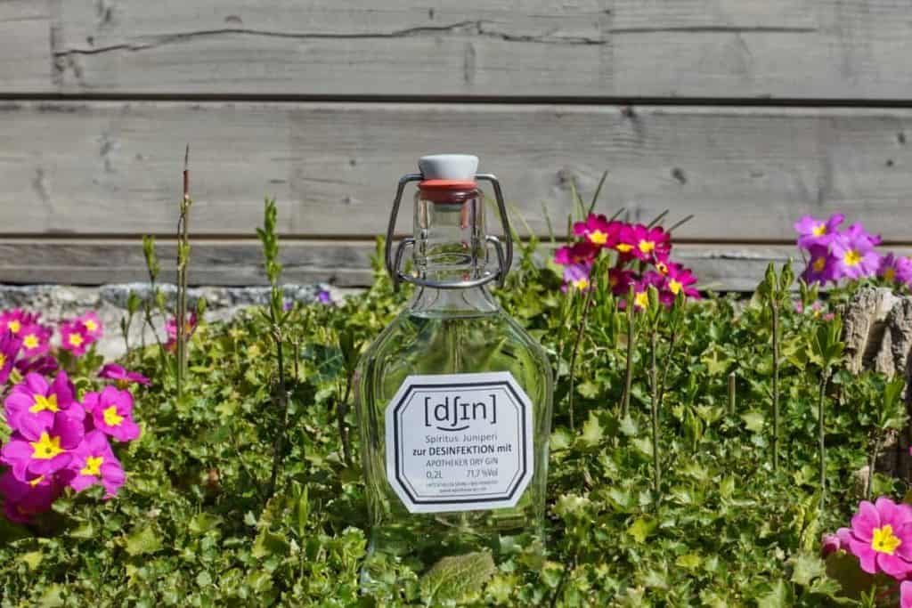 Eine durchsichtige Glasflasche mit einem Etikett steht auf einer Wiese, umgeben von rosa und gelben Blumen, im Hintergrund ist eine Holzwand zu sehen.