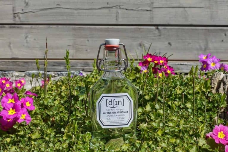 Eine durchsichtige Glasflasche mit einem Etikett steht auf einer Wiese, umgeben von rosa und gelben Blumen, im Hintergrund ist eine Holzwand zu sehen.