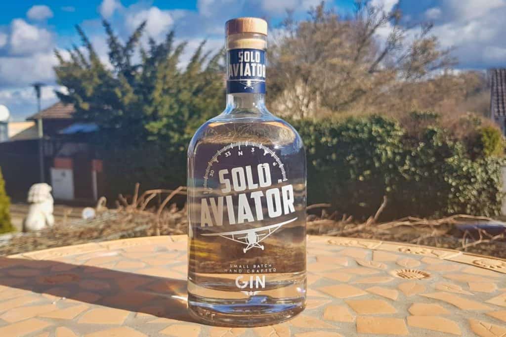Auf einem Mosaiktisch im Freien steht eine Flasche Solo Aviator Gin, im Hintergrund sind Bäume, Sträucher und ein teilweise bewölkter Himmel zu sehen.
