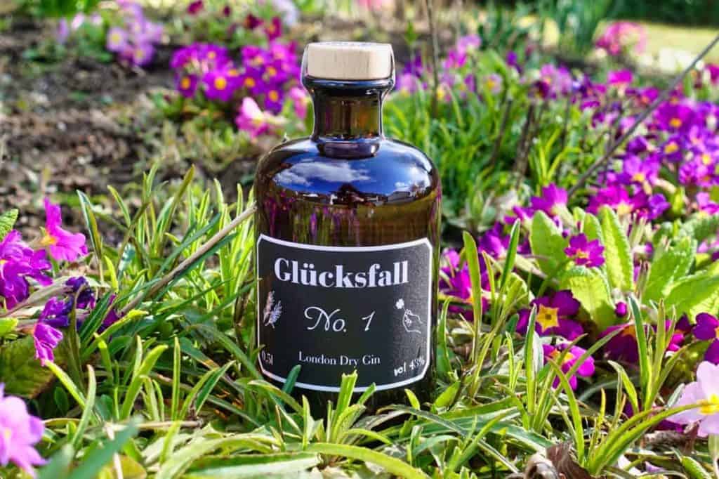 In einem Garten steht eine dunkle Glasflasche mit der Aufschrift „Glücksfall No. 1 London Dry Gin“ inmitten von leuchtend rosa und gelben Blumen und grünem Gras.