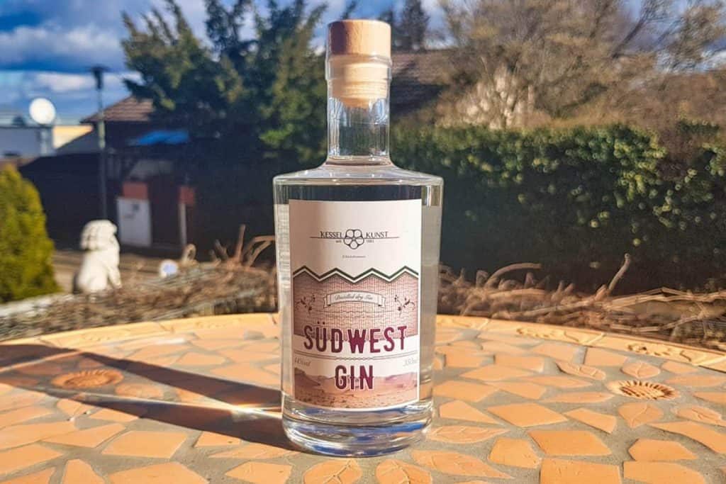 Auf einem mit Mosaikfliesen ausgelegten Tisch im Freien steht eine Flasche Südwest Gin, im Hintergrund sind Bäume, Büsche und Häuser zu sehen, über ihnen ein teilweise sonniger Himmel.