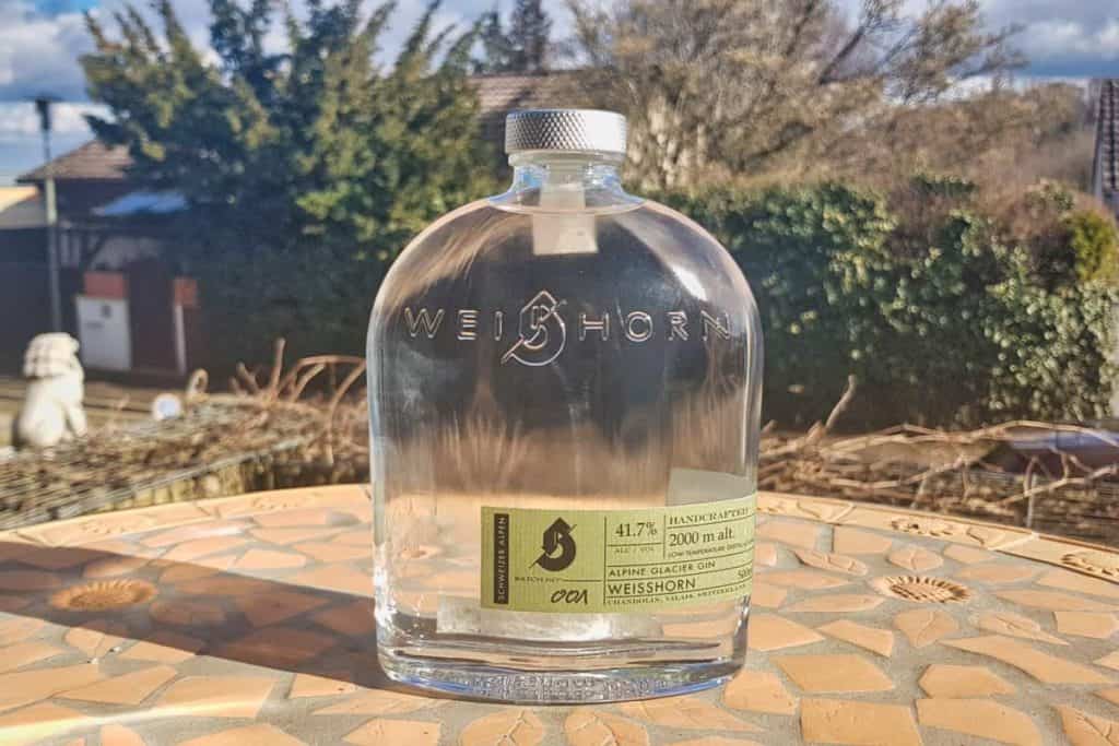 Auf einem Mosaiktisch im Freien steht eine durchsichtige Flasche „WEISSHORN Alpine Glacier Gin“, im Hintergrund sind Bäume, Dächer und ein blauer Himmel zu sehen.