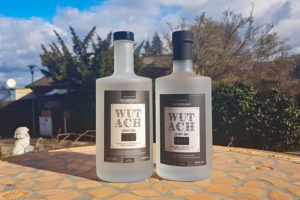 Zwei mattierte Flaschen „Wutach Japan Gin“ stehen auf einem sonnenbeschienenen Tisch im Freien, im Hintergrund sind Bäume und ein Haus vor einem teilweise bewölkten Himmel zu sehen.